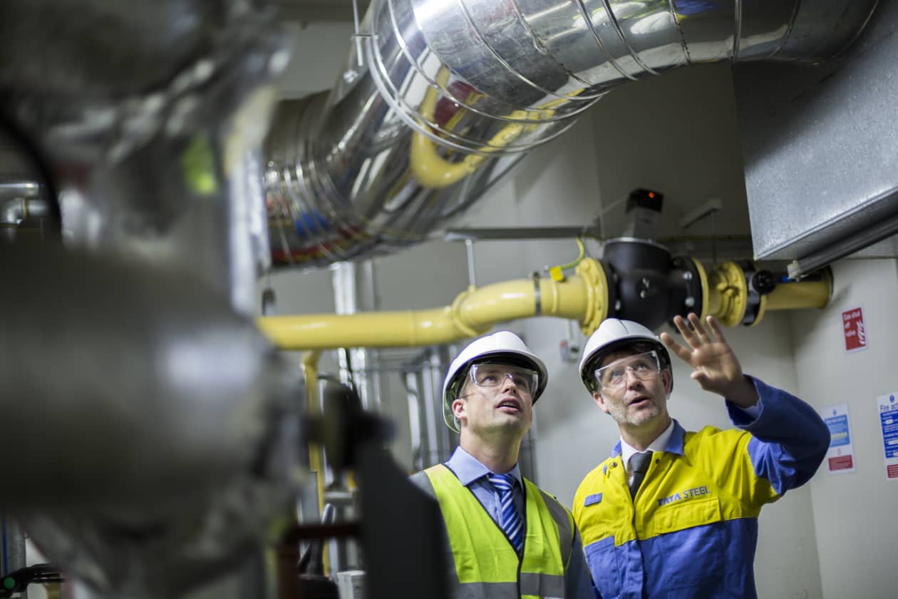 Two workers in safety gear and helmets inspect industrial pipes and valves.